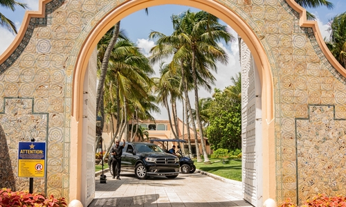 shutterstock_2476204587.jpg View through an ornate archway leading to a tropical entrance with palm trees and parked cars