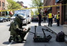 A bomb squad technician operating a robot in a street surrounded by police and emergency personnel