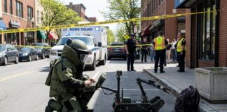 A bomb squad technician operating a robot in a street surrounded by police and emergency personnel