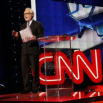 news anchor stands on stage beside a clear podium