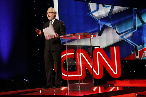 news anchor stands on stage beside a clear podium