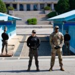 Two military personnel standing in front of blue buildings at a border
