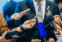 DOJ Weighs Perjury Probe After Senate Challenges Testimony Journalists holding microphones interview a person in a suit.