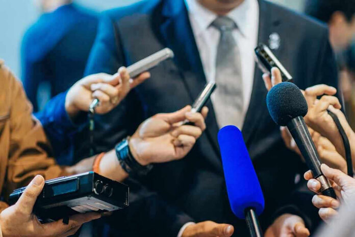 471987565-1 Journalists holding microphones interview a person in a suit.