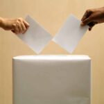 Two hands placing ballots into a voting box.